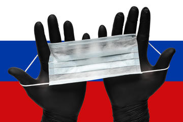 Two hands in black gloves hold a face mask with the Russian flag in the background